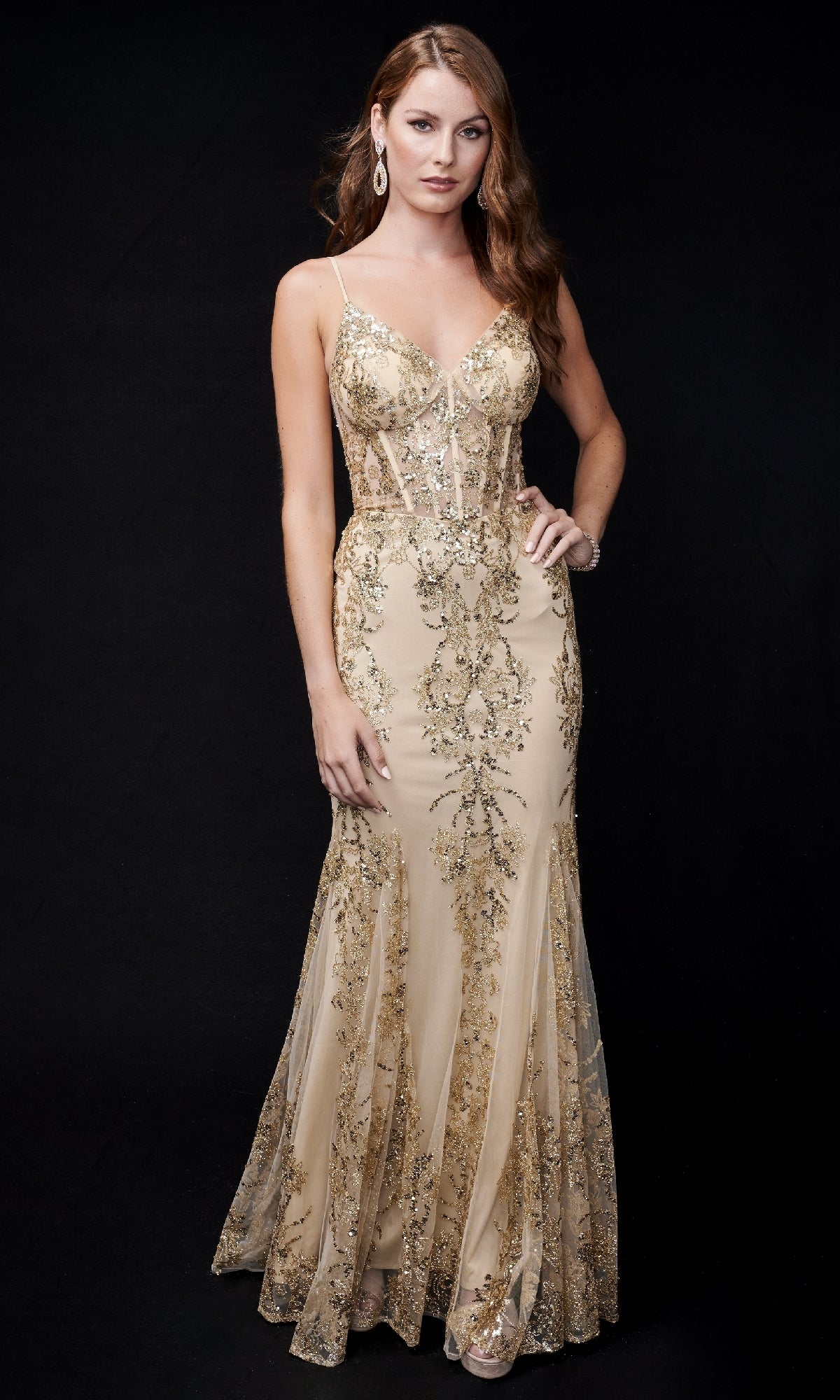 A woman wearing the Nina Canacci 1562 long formal dress, a sleeveless nude mermaid gown with gold floral embroidery, stands against a black background.